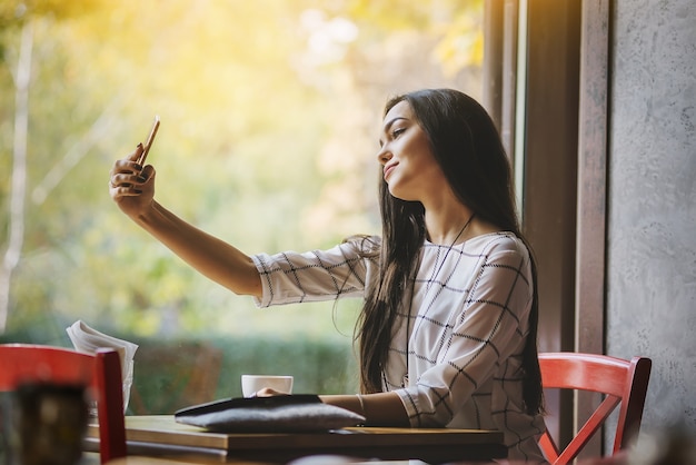 Jeune belle brune est assise dans un café et fait un téléphone selfie.