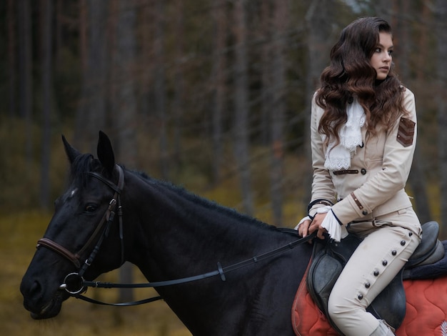 Une jeune belle cavalière brune dans un élégant costume rétro chevauchant un cheval noir dans une zone forestière