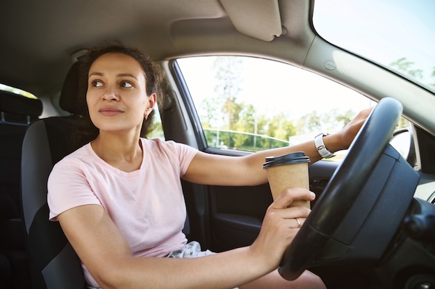 Jeune belle conductrice non attachée regardant sur le côté tout en conduisant une voiture et en buvant du café dans une tasse à emporter