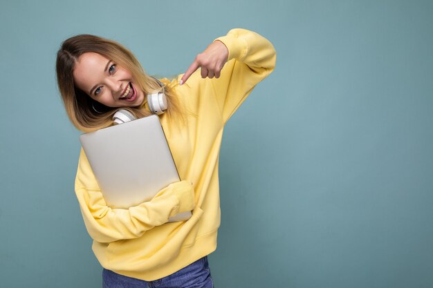 Jeune belle étudiante blonde portant un sweat à capuche jaune à la mode et des écouteurs tenant un ordinateur portable