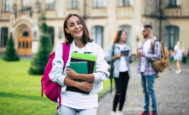 Jeune belle étudiante brune en vêtements en jean avec sac à dos et livres en mains sur un groupe de ses amis étudiants