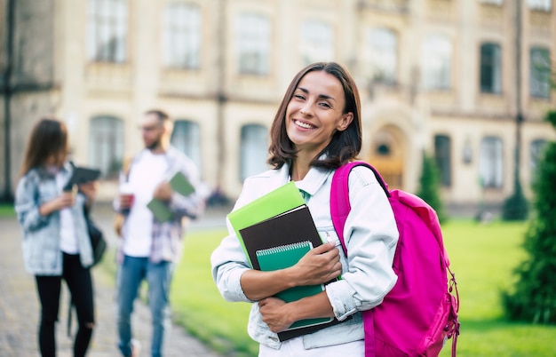 Jeune belle étudiante brune en vêtements en jean avec sac à dos et livres en mains sur un groupe de ses amis étudiants