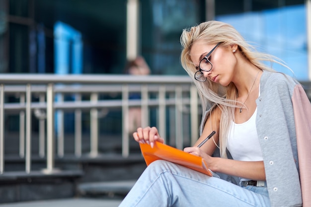 Jeune belle étudiante écrivant et lisant un livre dans les escaliers de l'université ou du collège, se préparant aux examens. Concept d'éducation, espace de copie.