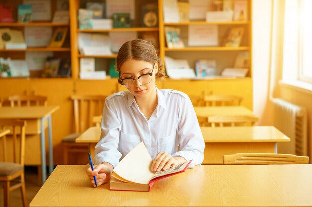 Jeune belle étudiante avec des lunettes, assise à une table dans le bureau et lisant un livre.