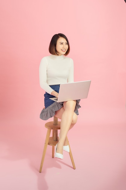 Jeune belle femme d'affaires asiatique utilisant un ordinateur portable assis sur une chaise. Isolé sur fond rose.