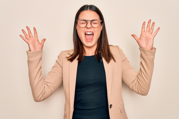 Jeune belle femme d'affaires brune portant une veste et des lunettes sur fond blanc célébrant la folie et la folie du succès avec les bras levés et les yeux fermés criant excité concept de gagnant