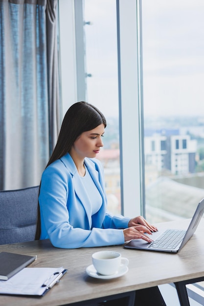 Jeune belle femme d'affaires dans une chemise bleue assise au bureau avec un ordinateur portable et faisant des affaires