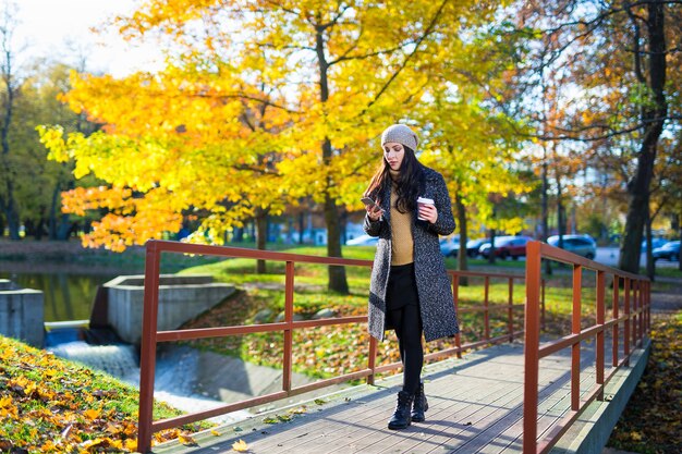 Jeune belle femme d'affaires avec un téléphone intelligent et une tasse de café marchant dans le parc d'automne