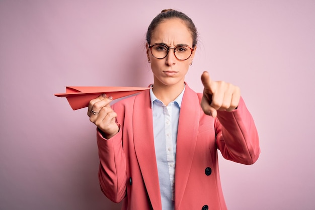 Jeune belle femme d'affaires tenant un avion en papier sur fond rose isolé pointant du doigt vers la caméra et vers vous signe de la main geste positif et confiant de l'avant