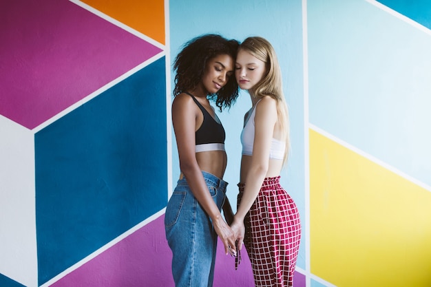 Jeune belle femme afro-américaine aux cheveux bouclés foncés et jolie femme aux cheveux blonds en hauts sportifs fermant les yeux rêveusement et se tenant la main avec un mur coloré