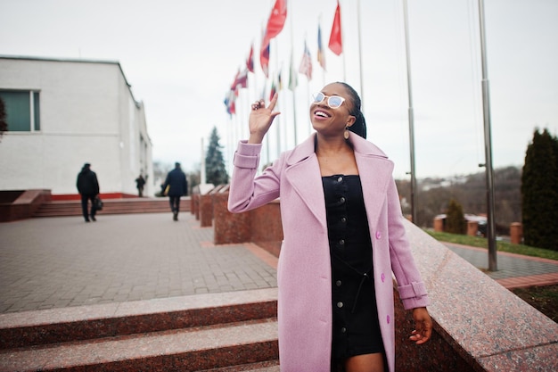 Jeune belle femme afro-américaine élégante dans la rue portant un manteau de tenue de mode et des lunettes contre les drapeaux de différents pays du monde