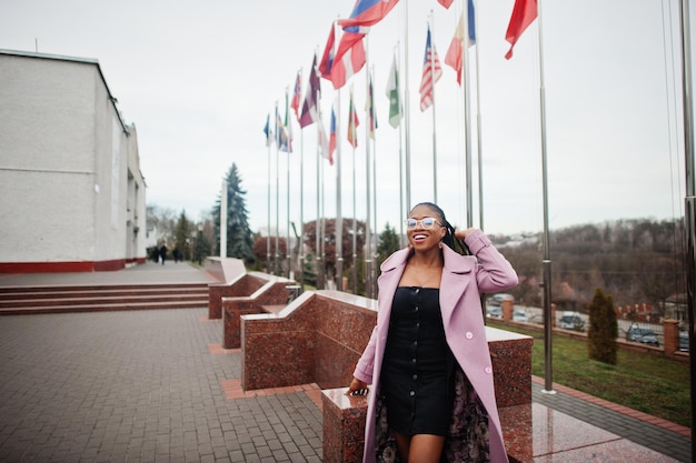 Jeune belle femme afro-américaine élégante dans la rue portant un manteau de tenue de mode et des lunettes contre les drapeaux de différents pays du monde