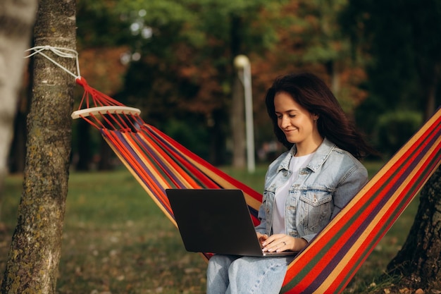 Jeune belle femme allongée dans un hamac avec un ordinateur portable dans un jardin fond vert arbres télétravail travail à distance photo de haute qualité