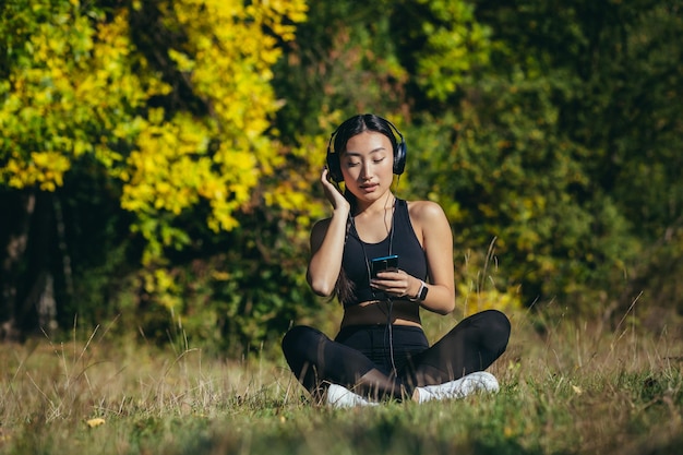 Jeune belle femme asiatique assise en position du lotus dans le parc, se relaxant après la forme physique et la course, écoutant de la musique sur des écouteurs et utilisant une application mobile sur le téléphone pour la forme physique