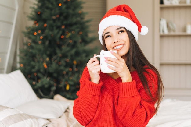Jeune belle femme asiatique aux cheveux longs noirs en pull rouge confortable et bonnet de Noel s'amusant avec une tasse de cacao sur le lit dans la chambre avec sapin de Noël