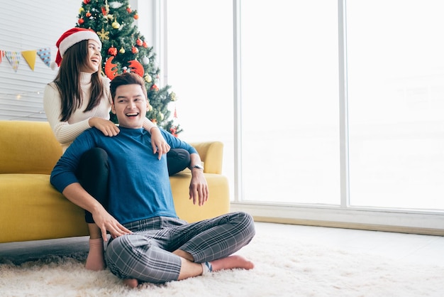 Jeune belle femme asiatique heureuse portant un chapeau de père Noël surprend et rit dans le dos de son petit ami à la maison avec un arbre de Noël en arrière-plan Image avec espace de copie