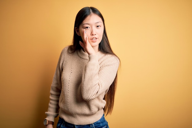 Jeune belle femme asiatique portant un pull décontracté sur fond isolé jaune main sur la bouche racontant une rumeur secrète chuchotant une conversation malveillante