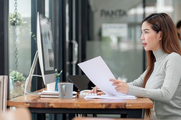 Jeune belle femme asiatique en pull de laine travaillant avec ordinateur et paperasse au bureau, vue latérale.