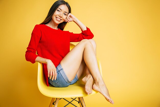 Jeune belle femme asiatique souriante à la mode en rouge est assise sur une chaise et possing isolé sur fond jaune