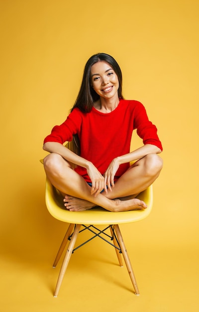 Jeune belle femme asiatique souriante à la mode en rouge est assise sur une chaise et possing isolé sur fond jaune