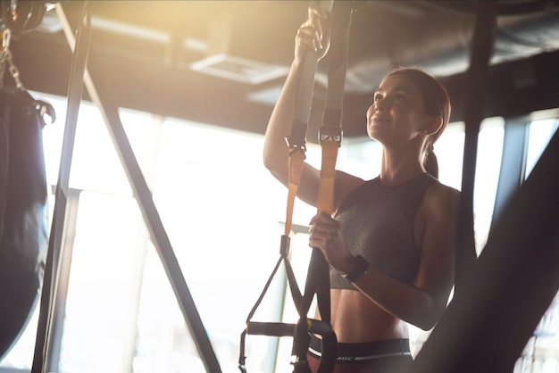 Jeune belle femme athlétique en vêtements de sport ajustant les sangles de fitness trx en se tenant debout au gymnase