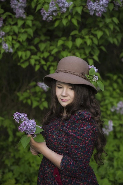 Jeune belle femme au chapeau se tient devant un buisson de lilas Couronne de lilas sur le chapeau