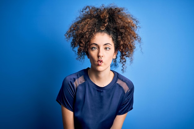 Jeune belle femme aux cheveux bouclés et piercing portant un t-shirt bleu décontracté faisant face au poisson avec des lèvres geste fou et comique Expression drôle