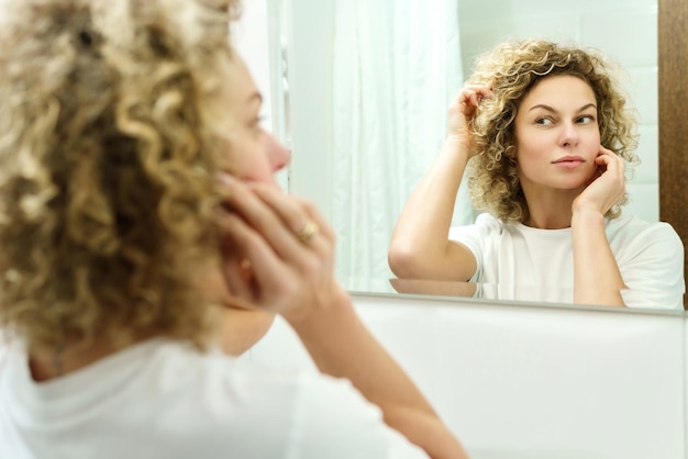 Jeune et belle femme aux cheveux bouclés regardant dans le miroir dans une salle de bain