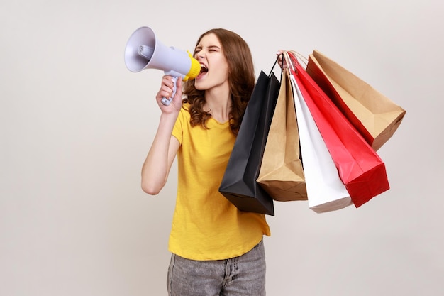 Jeune belle femme aux cheveux bruns ondulés porte un T-shirt jaune de style décontracté tenant des sacs à provisions et un mégaphone, annonçant la vente et la remise. Tourné en studio intérieur isolé sur fond gris.