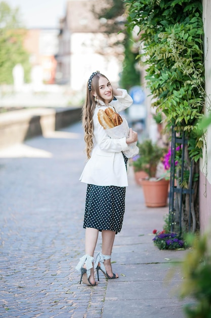 Jeune belle femme aux cheveux longs de 25 ans vêtue d'une robe noire et blanche achète une baguette française au centre d'une ville européenne Boulangerie fraîche Un modèle élégant se promène dans la ville