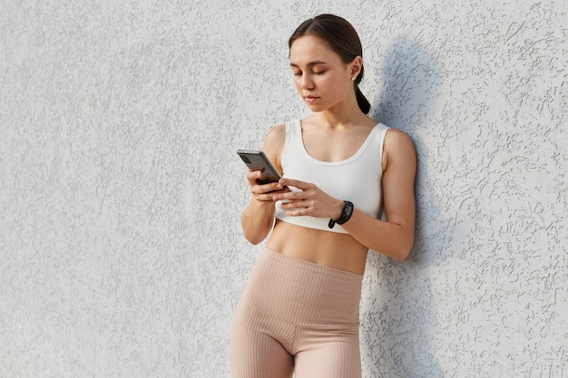 Jeune belle femme aux cheveux noirs portant un haut blanc et des leggins beiges, tenant et utilisant un téléphone intelligent dans les mains, debout près d'un mur gris, regardant l'écran du téléphone portable.