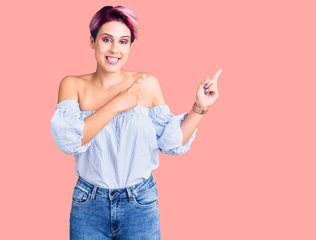 Jeune belle femme aux cheveux roses portant des vêtements décontractés souriant et regardant la caméra pointant avec deux mains et doigts sur le côté