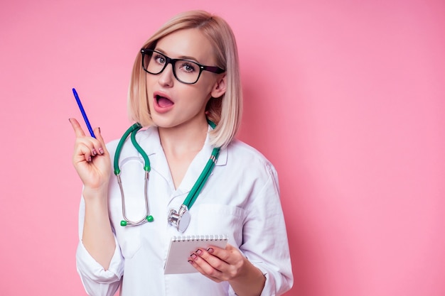 Jeune et belle femme blonde de Cupidon dans une blouse médicale blanche avec stéthoscope sur fond rose dans le studio. concept de jeux de rôle Saint Valentin Le 14 février