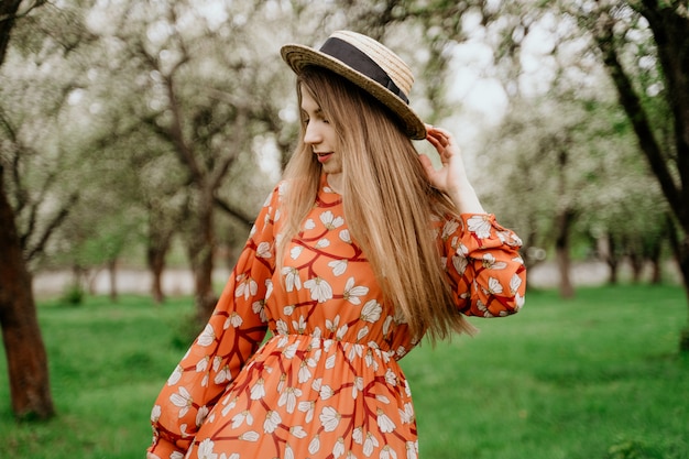 Jeune belle femme blonde dans un jardin fleuri. Arbres de printemps en fleurs. Robe orange et chapeau de paille.