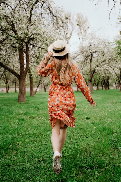 Jeune belle femme blonde dans un jardin fleuri. Arbres de printemps en fleurs. Robe orange et chapeau de paille.