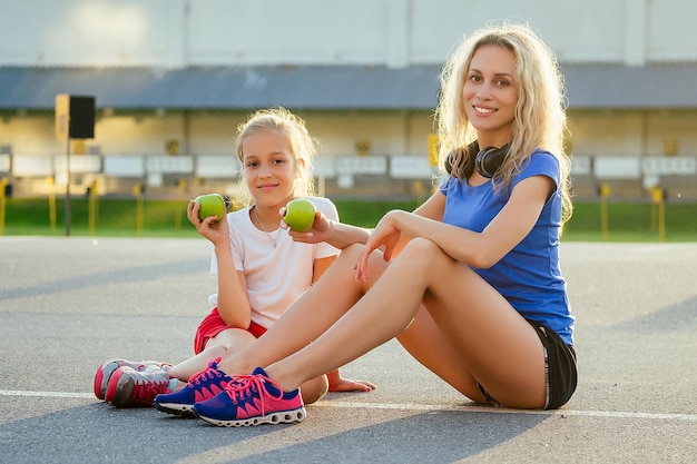Jeune belle femme blonde avec une petite fille en tenue de sport assise sur l'asphalte et mangeant une pomme verte au stade. Mère et fille de famille actives faisant du jogging le matin d'été