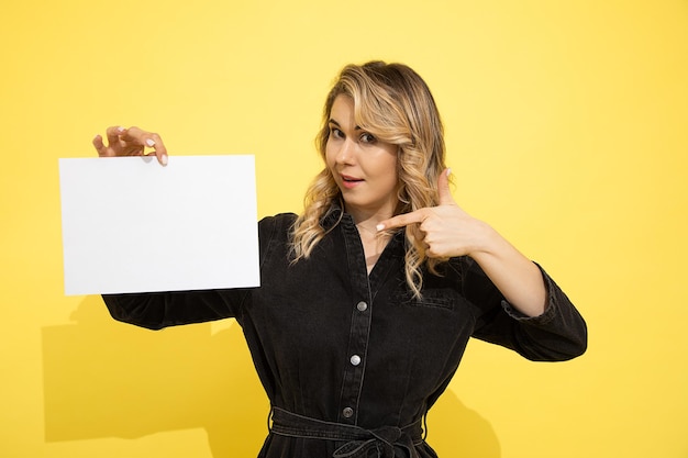 Jeune belle femme blonde en robe noire tient une feuille vide pour l'inscription sur fond jaune dans les mains pointant avec son doigt, sourit et rit joyeusement sur fond jaune en studio