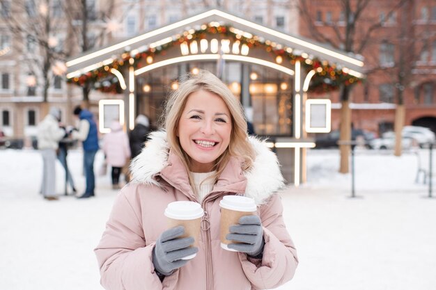 Jeune belle femme blonde souriante portant un manteau debout au-dessus des décorations de la ville de Noël et des lumières incandescentes tenir des tasses de café en plein air saison des fêtes de salutation d'hiver