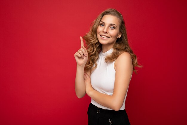 Jeune belle femme bouclée blonde positive souriante avec des émotions sincères portant un haut blanc isolé sur fond rouge avec un espace libre et pointant vers l'espace de copie pour le texte.