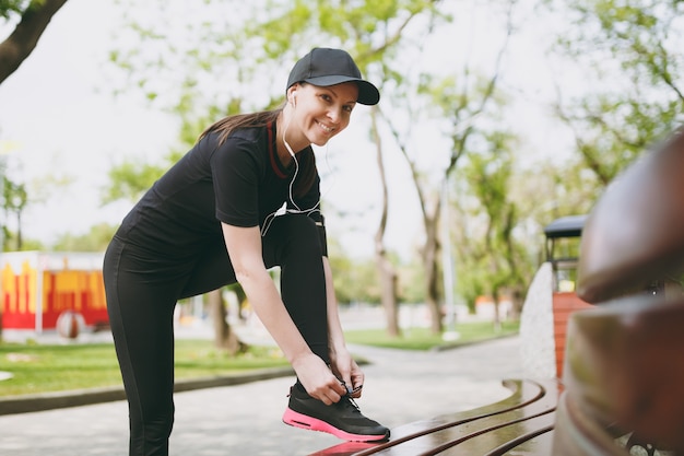 Jeune Belle Femme Brune Athlétique En Uniforme Noir Et Casquette Avec écouteurs écoutant De La Musique, Attachant Des Lacets Avant De Courir, S'entraînant Sur Un Banc Dans Le Parc De La Ville à L'extérieur