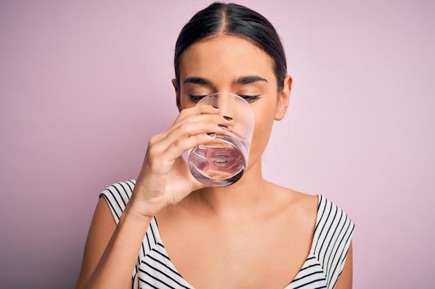 Jeune belle femme brune buvant un verre d'eau saine pour se rafraîchir debout sur fond rose isolé