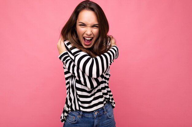 Jeune belle femme brune. Dame à la mode en manches longues à rayures décontractées. Une femme positive montre des émotions faciales sincères. Modèle drôle isolé sur fond rose avec espace libre.