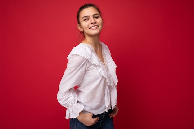 Jeune belle femme brune élégante européenne portant un chemisier blanc isolé sur fond rouge avec des émotions sincères positives. Simple et naturel en regardant la caméra. Espace libre.