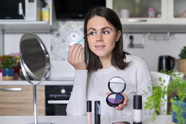 Jeune belle femme brune faisant le maquillage quotidien du visage avec des cosmétiques décoratifs et un miroir assis à la maison dans la cuisine, applique du fard à paupières