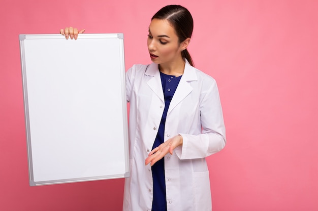 Jeune de belle femme brune heureuse portant un blouse blanche médicale tenant un tableau magnétique blanc pour maquette isolé sur fond rose avec un espace vide.