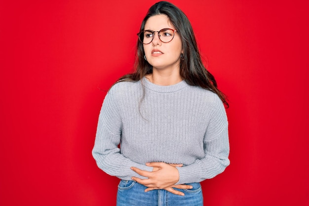 Jeune belle femme brune portant un pull décontracté sur fond rouge avec la main sur l'estomac parce que l'indigestion maladie douloureuse se sent malade concept de douleur