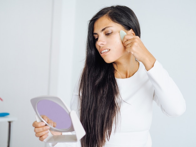 Jeune belle femme brune se faisant un massage du visage avec l'aide de la pierre de gua sha