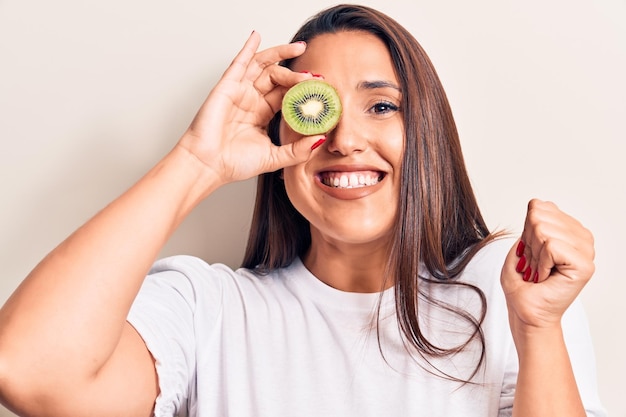 Jeune belle femme brune tenant un kiwi criant fier de célébrer la victoire et le succès très excité avec le bras levé