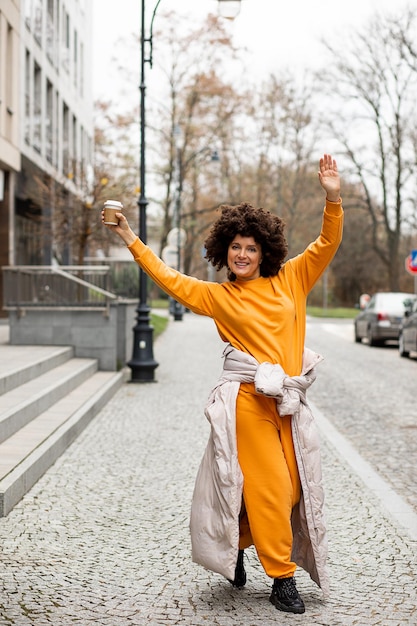 Photo jeune, belle femme caucasienne de 40 ans, vêtements de sport décontractés, dansant dans la rue avec une tasse de café jetable. coiffure afro. profiter de la vie le jour de l'automne.