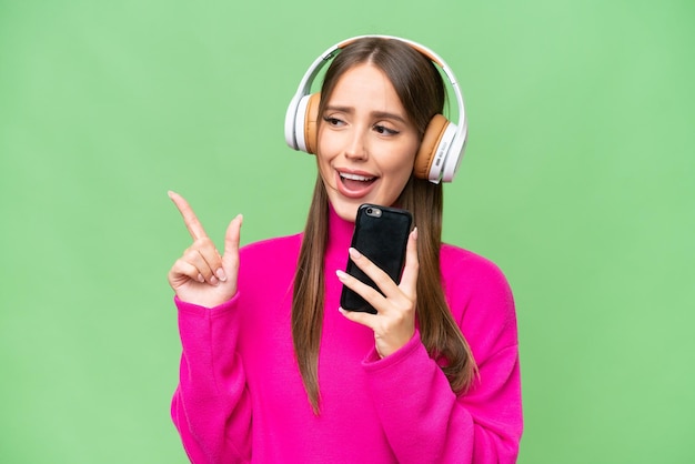 Jeune belle femme caucasienne sur fond isolé écoutant de la musique avec un mobile et chantant